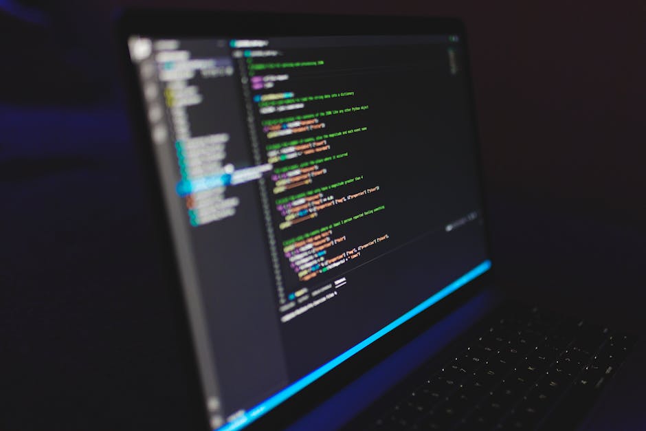 A close-up view of a laptop screen displaying lines of colorful code on a dark background, with a blurred dark environment in the background. The code includes various syntax elements highlighted in m